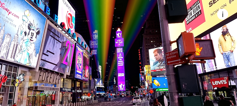 A rainbow made of light stretches across the sky over Times Square at night. 
