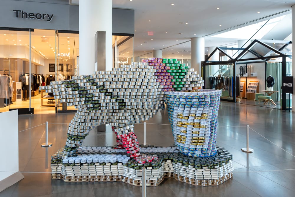 A sculpture made out of canned foods that depicts a pigeon drinking our of a Greek coffee cup