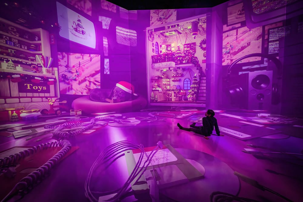 A person stands Two people sits in a room with walls covered in digital projections of a room filled with toys and holiday decorations