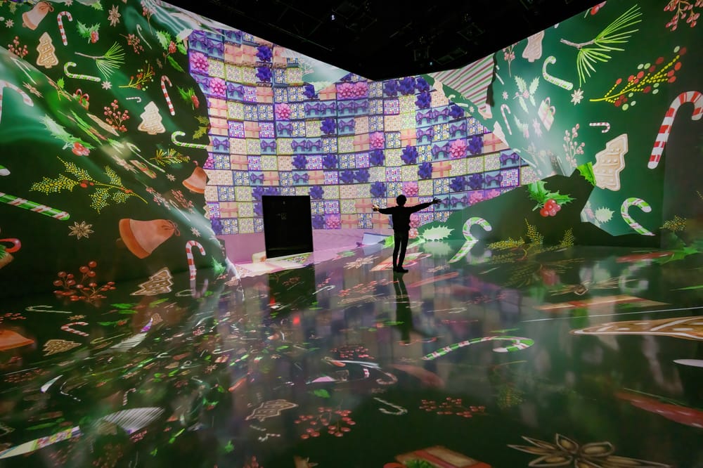 A person stands Two people stand in a room with walls covered in digital projections of holiday warpping paper