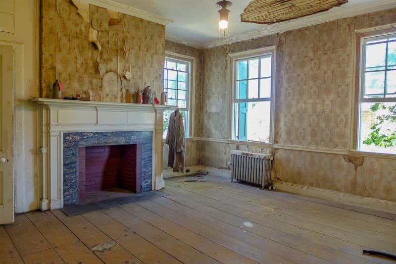 An empty room with a fireplace, radiator, three windows, and peeling beige wallpaper