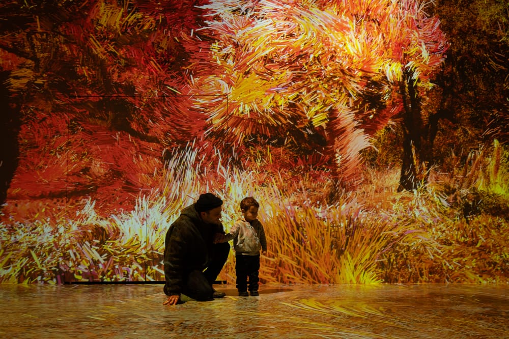 A small boy and a man are surrounded by abstract orange and yellow figures projected on the wall behind them and floor