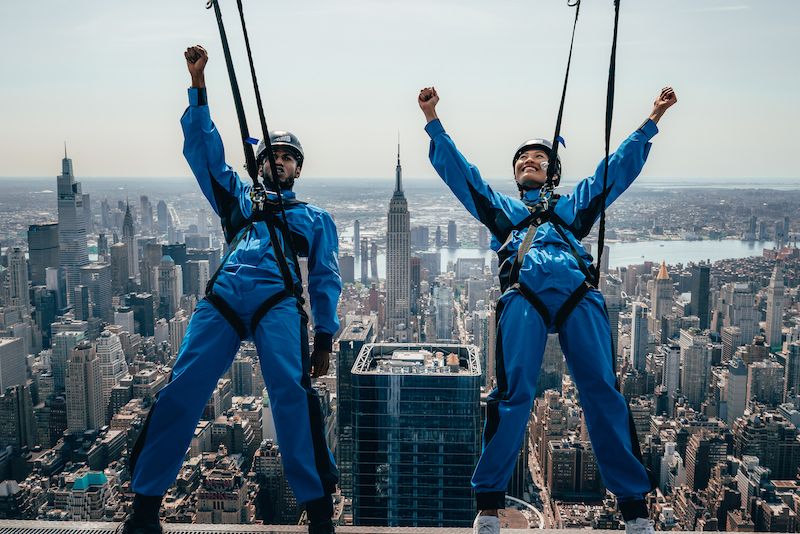 Two people hang off the side of a building with views of Manhattan