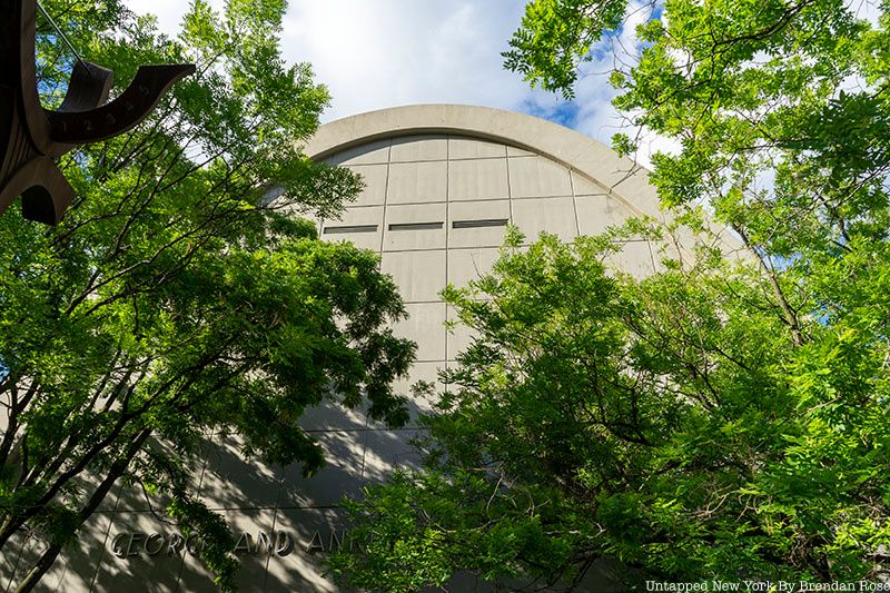 The arch of Asphalt Green seen through the trees, June 2022. A windowless building.