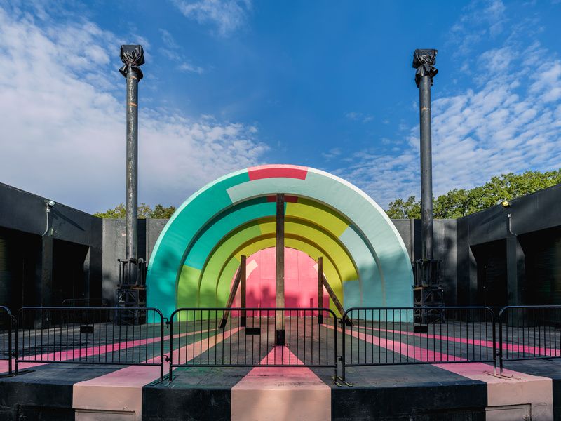 A pink, green, and blue mural covers the bandshell with wooden pillars on the stage.