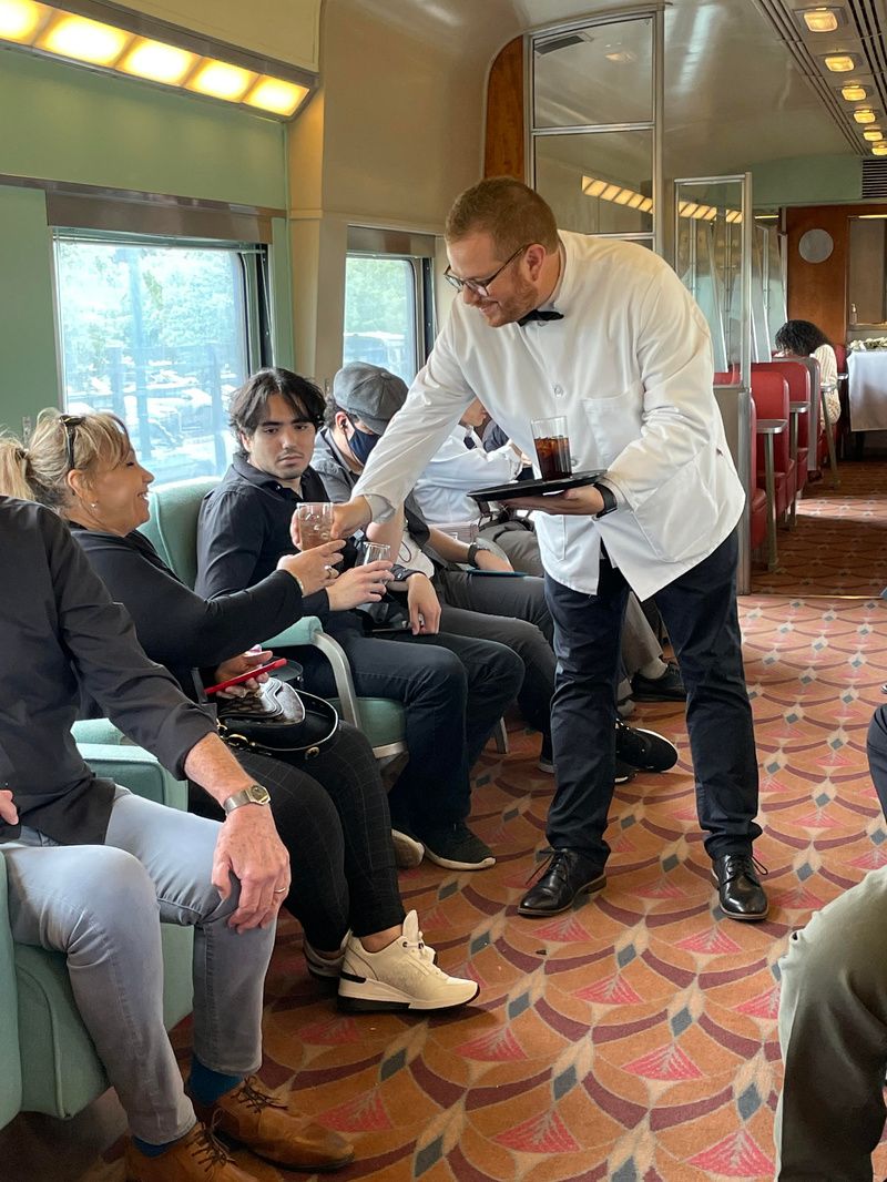 A waiter in a white jacket serves a drink to a women sitting inside a vintage train car