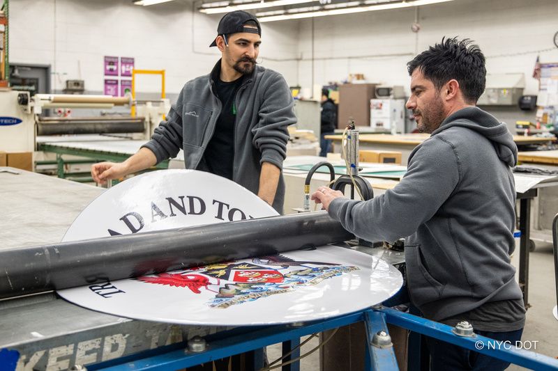 Two workers flatten out a country medallion for the Avenue of the Americas inside DOT's Sign Shop