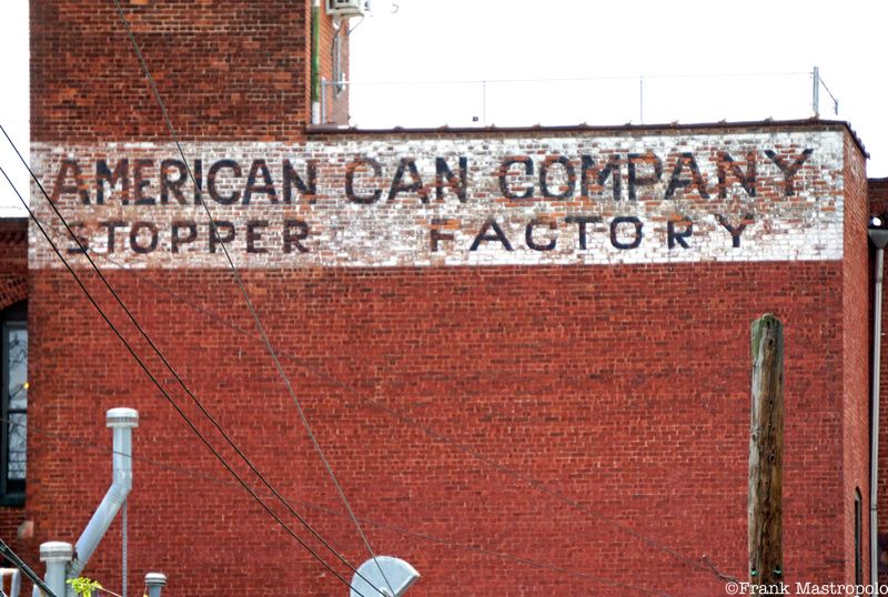 A painted sign with a white background that reads "American Can Company Stopper Factory" on a red brick wall