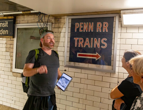 Untapped New York Guide Justin Rivers talking to tour guests in front of vintage sign inside Penn Station
