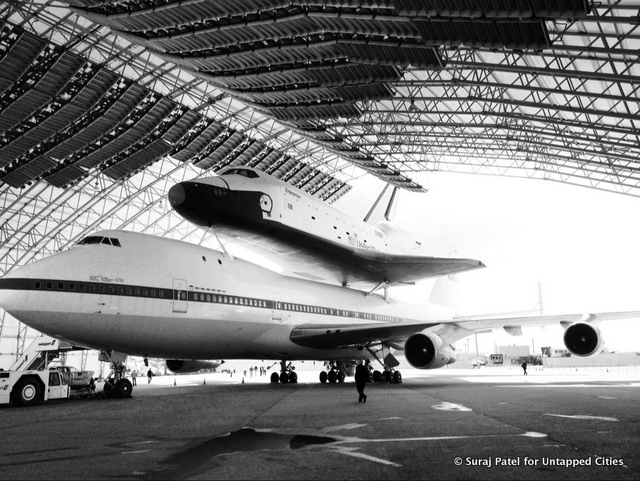 A space shuttle sits atop a plane inside a hangar