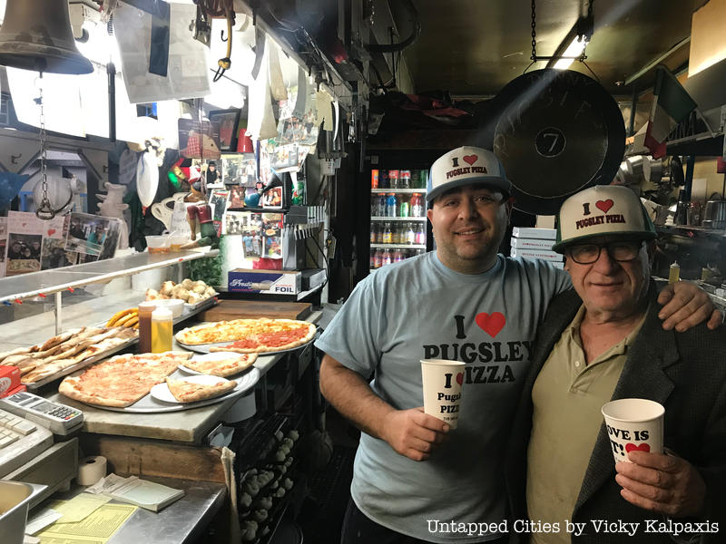 Two men stand next to a pizza counter at Pugsley Pizza