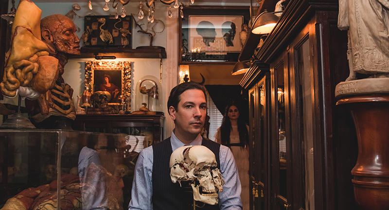 A man sits among a room of curiosities holding a skull