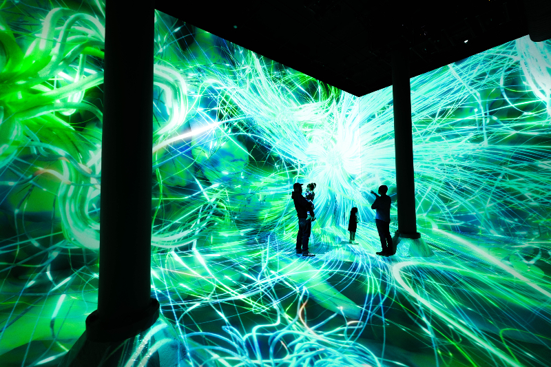 Adults and young people stand inside a digital exhibition surrounded by green and blue lights crossing the room.