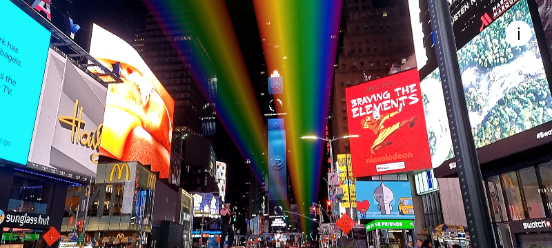 A rainbow made of light stretches across the sky over Times Square at night. 