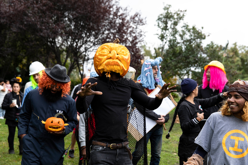 A man with a carved pumpkin on his head walks through a crowd of people in Halloween costumes at Socrates Sculpture Park.