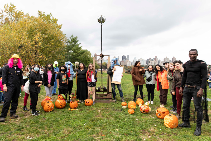 A group of people stand in front of their carved pumpkins and next to a large catapult waiting to send their jack-o-lanterns into the air.