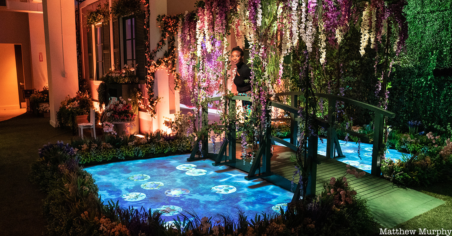 A woman stands on a flower covered bridge over looking a Monet painting projected on the floor at Monet's Garden