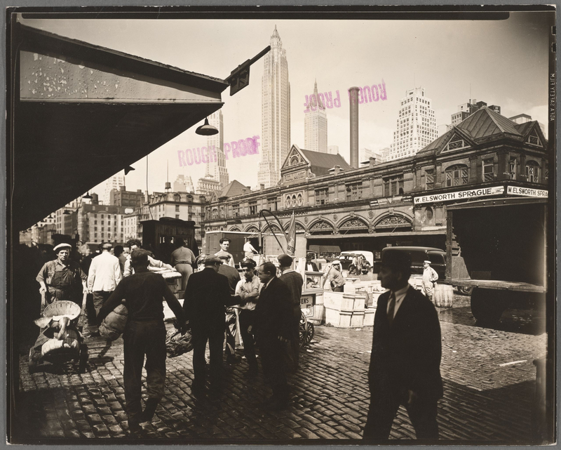 Black and white photo of the Fulton Fish Market in the 1930s with workers and people walking in front of it on the cobblestone streets and a few skyscrapers in the background
