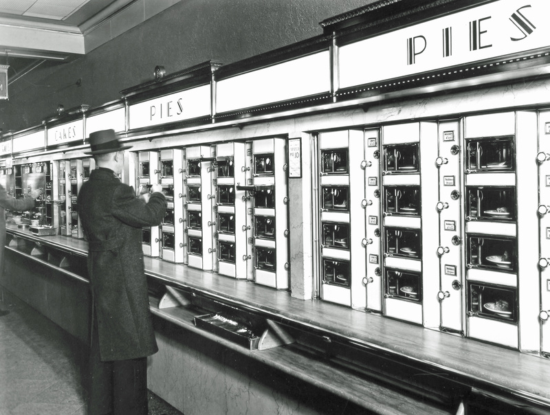 Black and white vintage photo of a man in a dark coat and hat ordering food at an automat in NYC