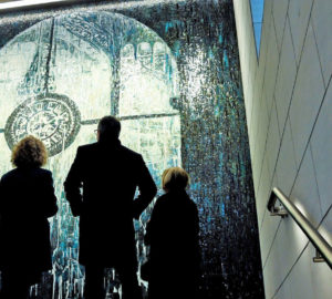 Three people stand in front of a new mosaic in Penn Station that depicts a clock framed by an arched window