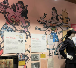 A mannequin in Wonder Woman's Army nurse uniform stands in front of a wall covered in framed images at the City Reliquary Wonder Woman exhibit