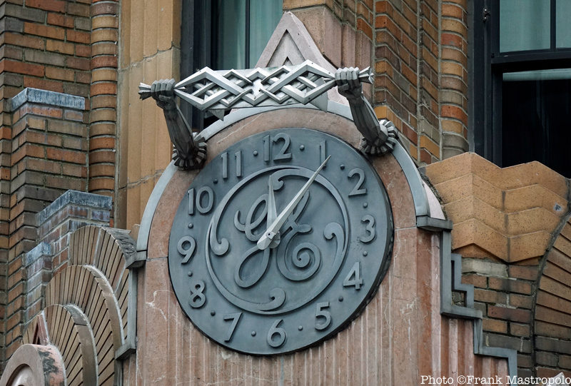 A gret clock with the GE logo in the middle under two arms holding silver radio waves at the top of the RCA Building