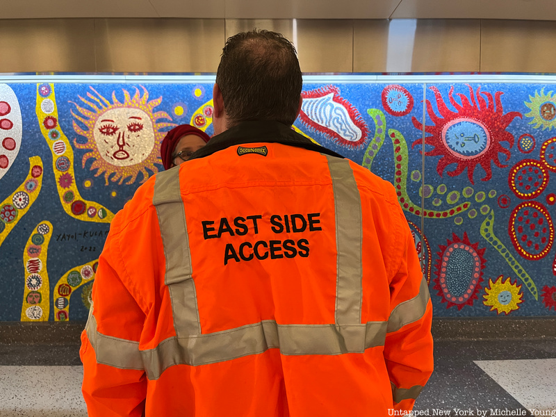 MTA worker in a bright orange jacket that reads "East Side Access" stands in front of a mosaic mural in Grand Central Madison