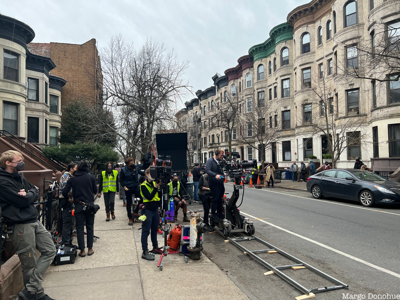 A film crew moves a camera along dolly track in front of a street of Brooklyn rowhouses