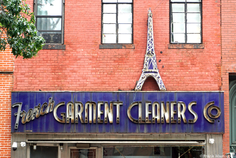 Front of a brick building with a blue rectangular Brooklyn ghost sign that reads "French Garment Cleaners Co." and the shape of the Eiffel Tower