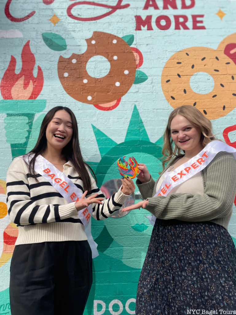 Two women hold a rainbow bagel in front of a bagel mural