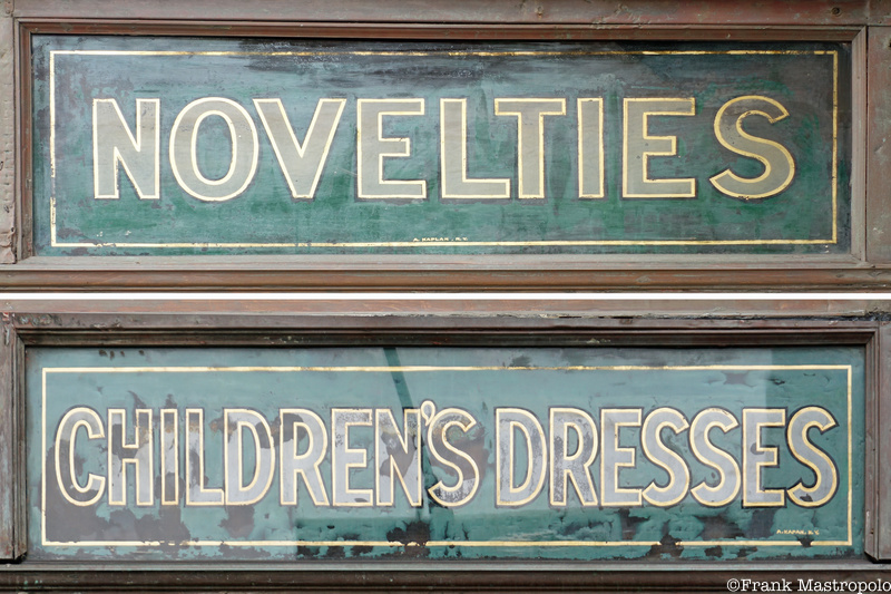A green sign with beige letters that reads "Novelties" and "Children's Dresses"