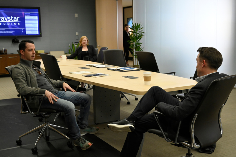 Three people sit at a conference table in a meeting room in a scene in Succession