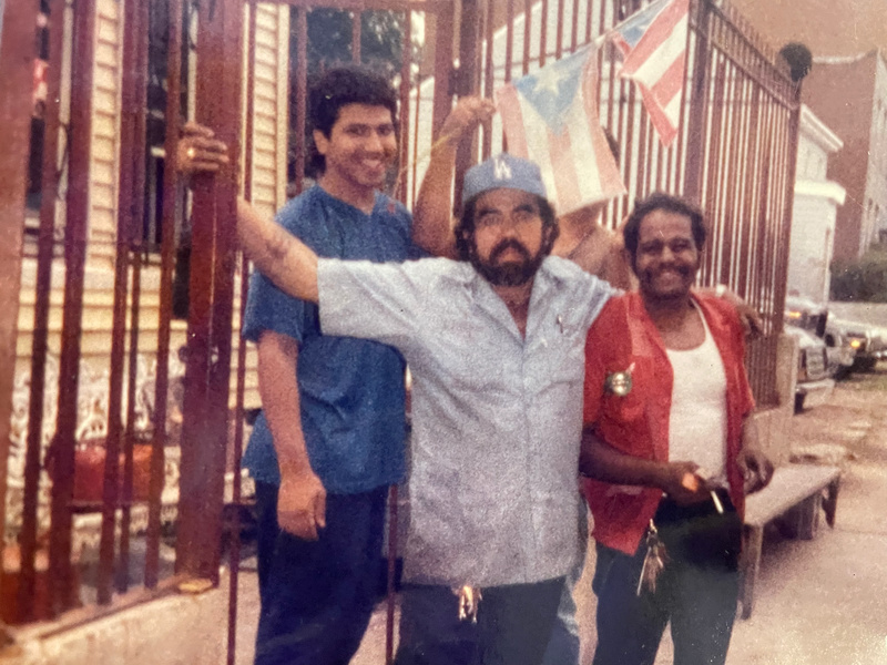 Three men stand in front of a fence and Puerto Rican flags