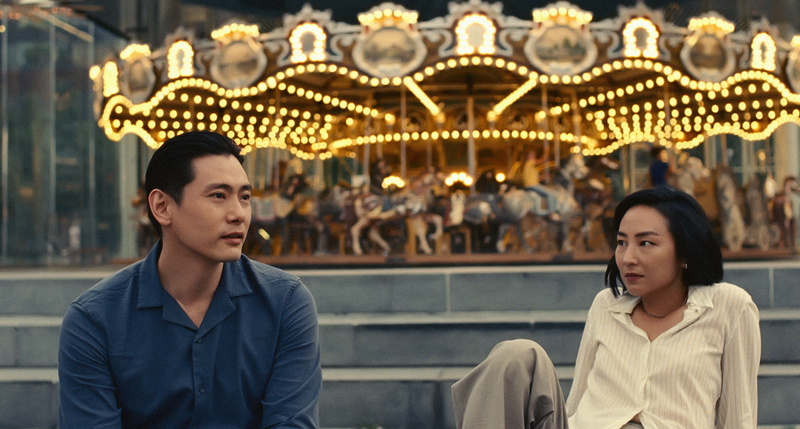 Teo Yoo and Greta Lee in front of a carousel